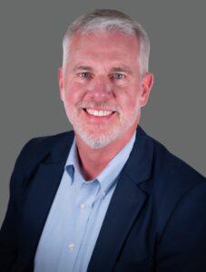 Man with gray hair and beard smiling in a blue suit jacket and light colored shirt against a gray background.