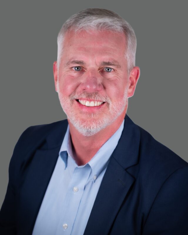Man with gray hair and beard smiling in a blue suit jacket and light colored shirt against a gray background.