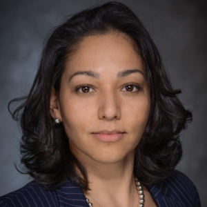 Portrait of a woman with shoulder-length dark hair, wearing a pinstripe suit and necklace, against a blurred gray background.