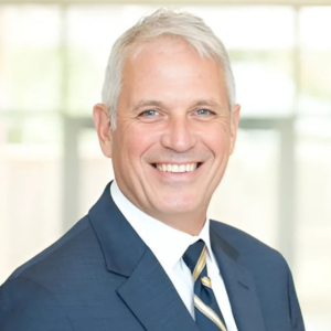 Smiling man in a suit and tie with short gray hair standing in a bright room.