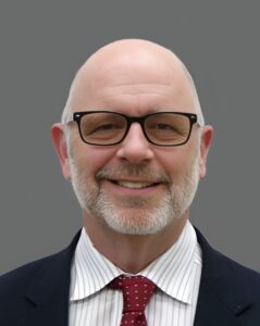Bald man with glasses and a beard wearing a suit, striped shirt, and polka dot tie, smiling against a gray background.