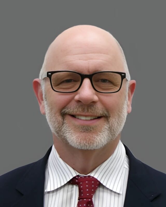 Bald man with glasses and a beard wearing a suit, striped shirt, and polka dot tie, smiling against a gray background.