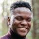 Smiling person wearing a maroon turtleneck in a forest background.