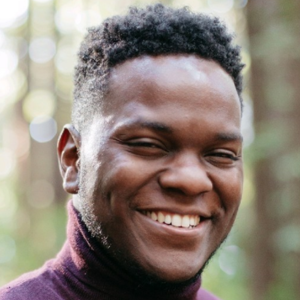 Smiling person wearing a maroon turtleneck in a forest background.