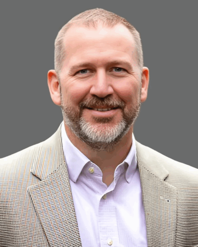 Smiling man in a suit jacket and shirt, standing against a gray background.