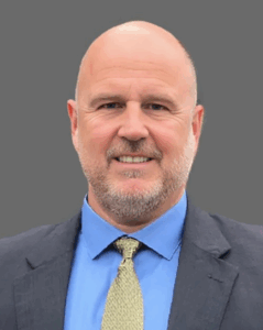 Man in a suit and tie smiling against a gray background.