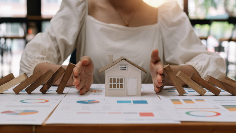 Person protecting a small house model from falling dominoes on top of financial documents in a bright room.