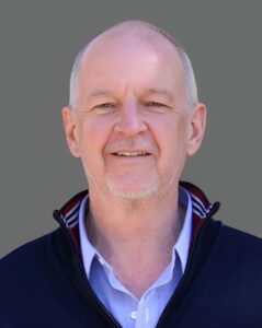 Smiling man with a bald head, wearing a black jacket and light blue shirt, standing against a gray background.