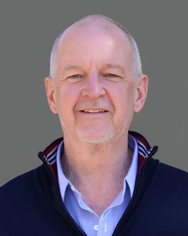 Smiling man with a bald head, wearing a black jacket and light blue shirt, standing against a gray background.