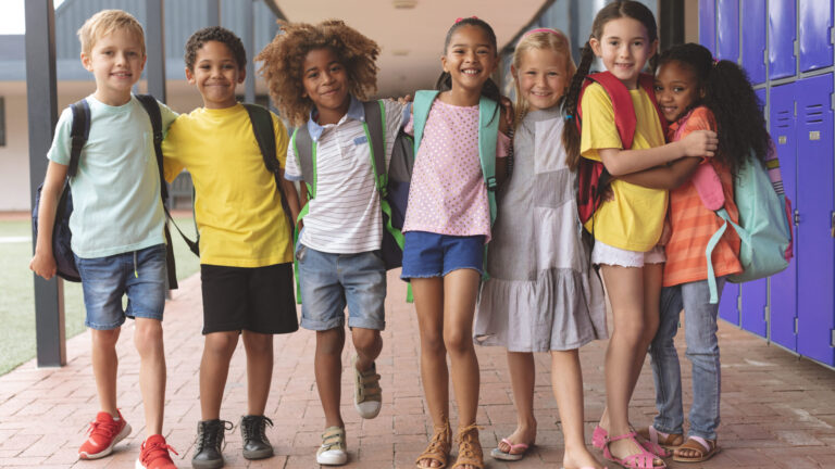 Group of smiling children with backpacks standing in school corridor near lockers.