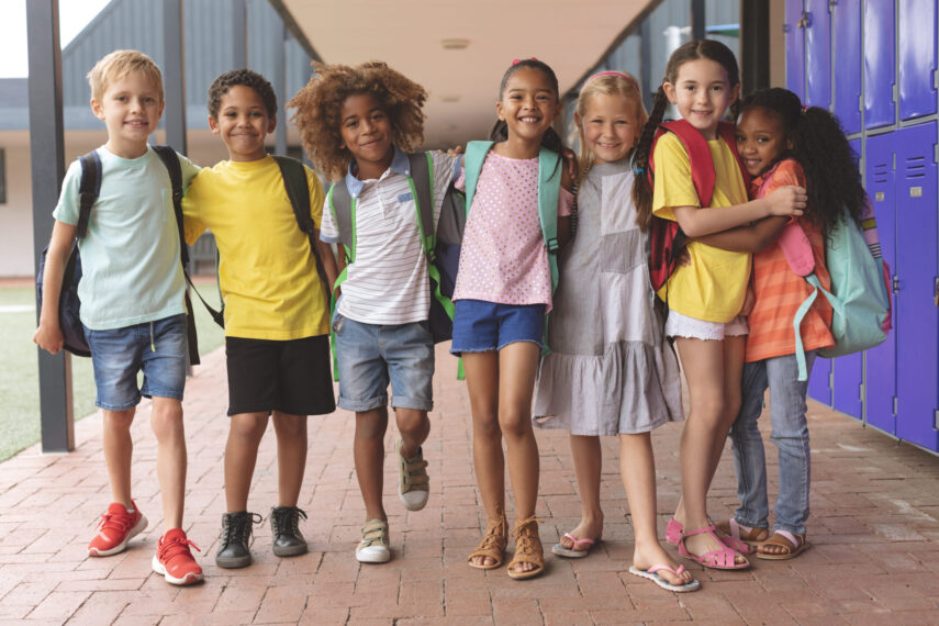 Group of smiling children with backpacks standing in school corridor near lockers.