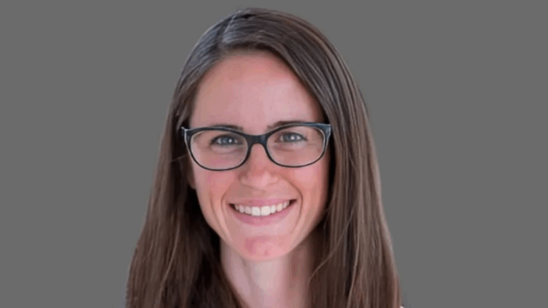 Smiling person with long brown hair and glasses against a gray background.