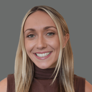 Smiling person with long blonde hair wearing a brown sleeveless top against a gray background.