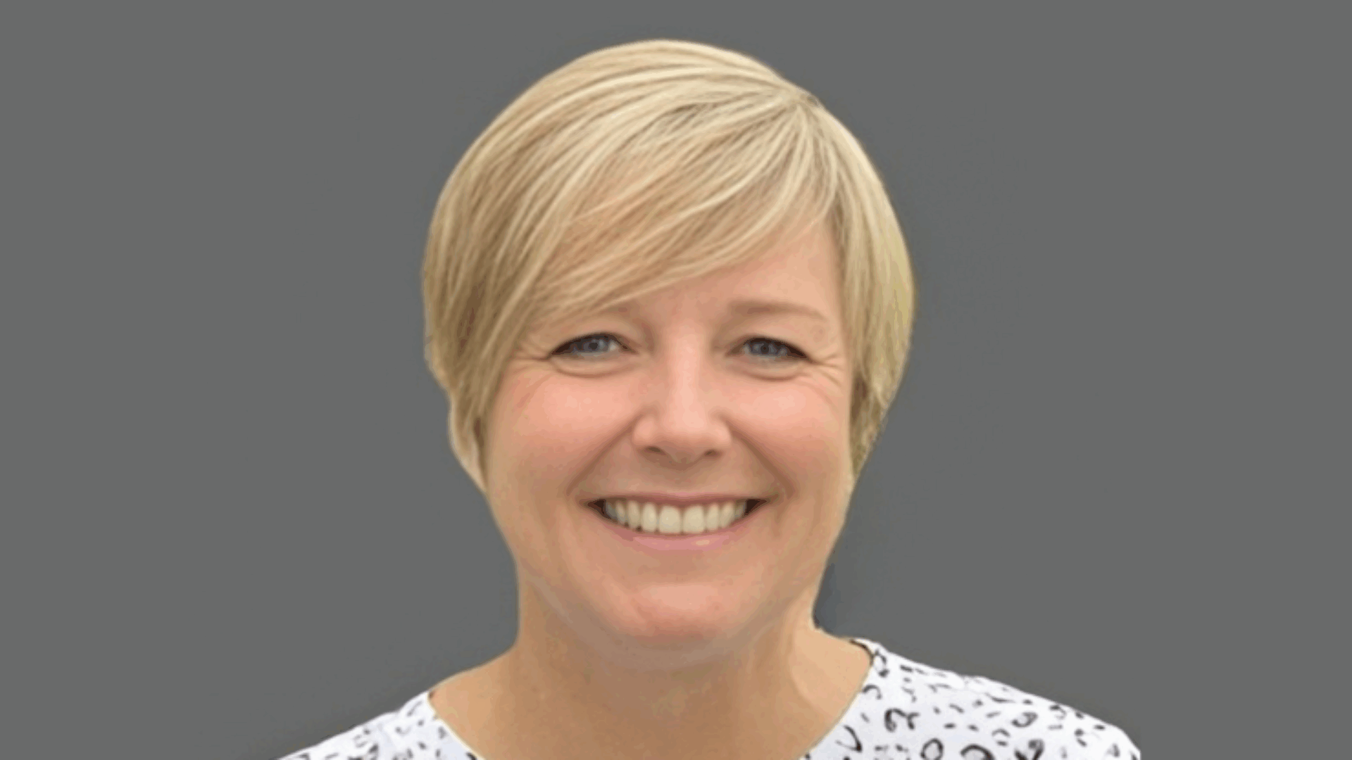 Person with short blonde hair smiling against a gray background wearing a patterned shirt.