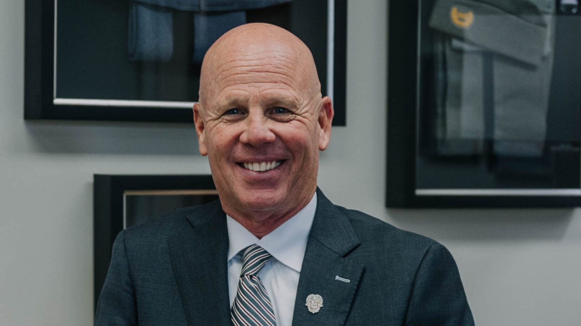 Man in a suit leaning on a wooden desk, smiling, with framed pictures in the background.