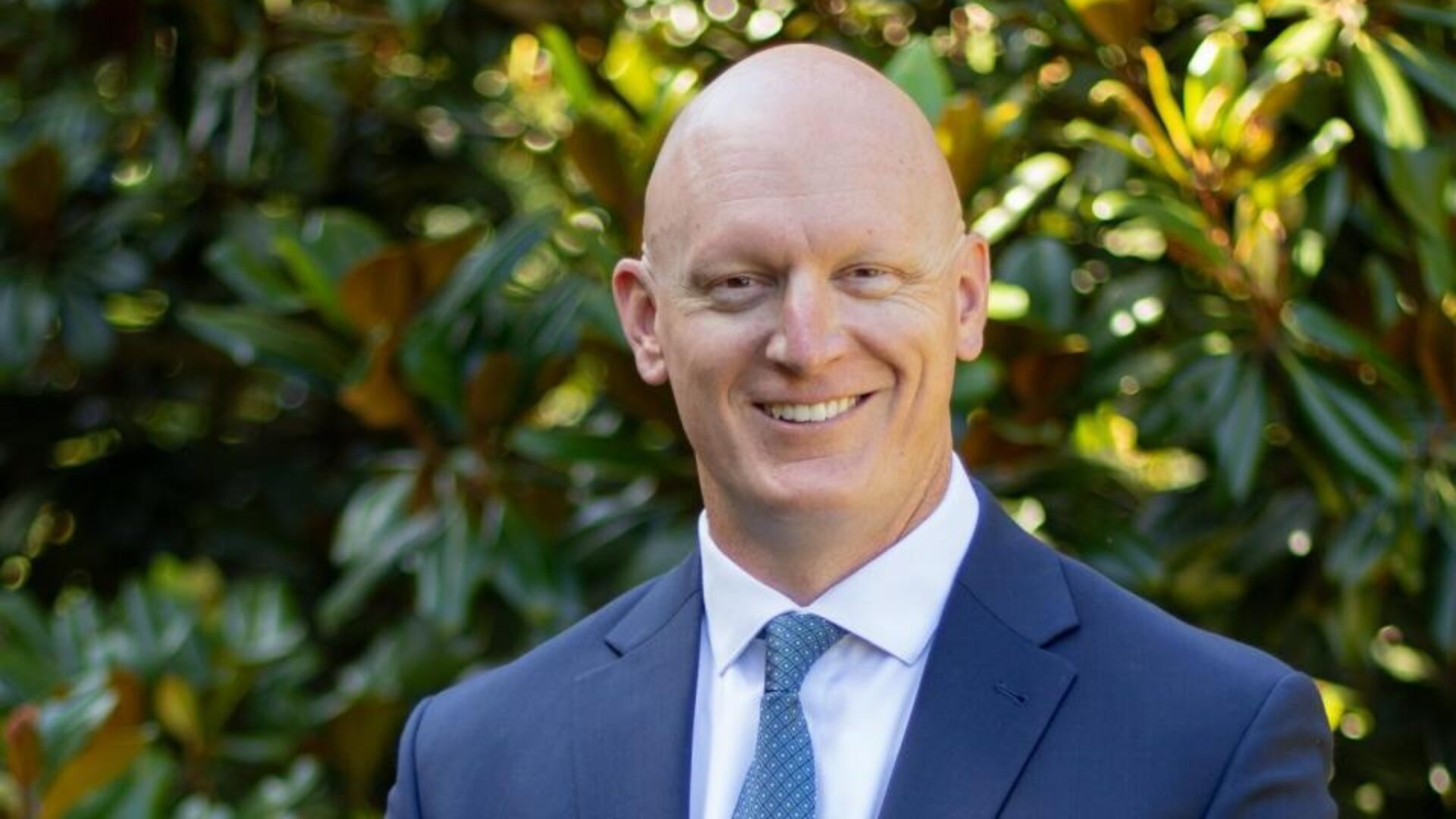 Smiling man in a suit standing outdoors in front of greenery