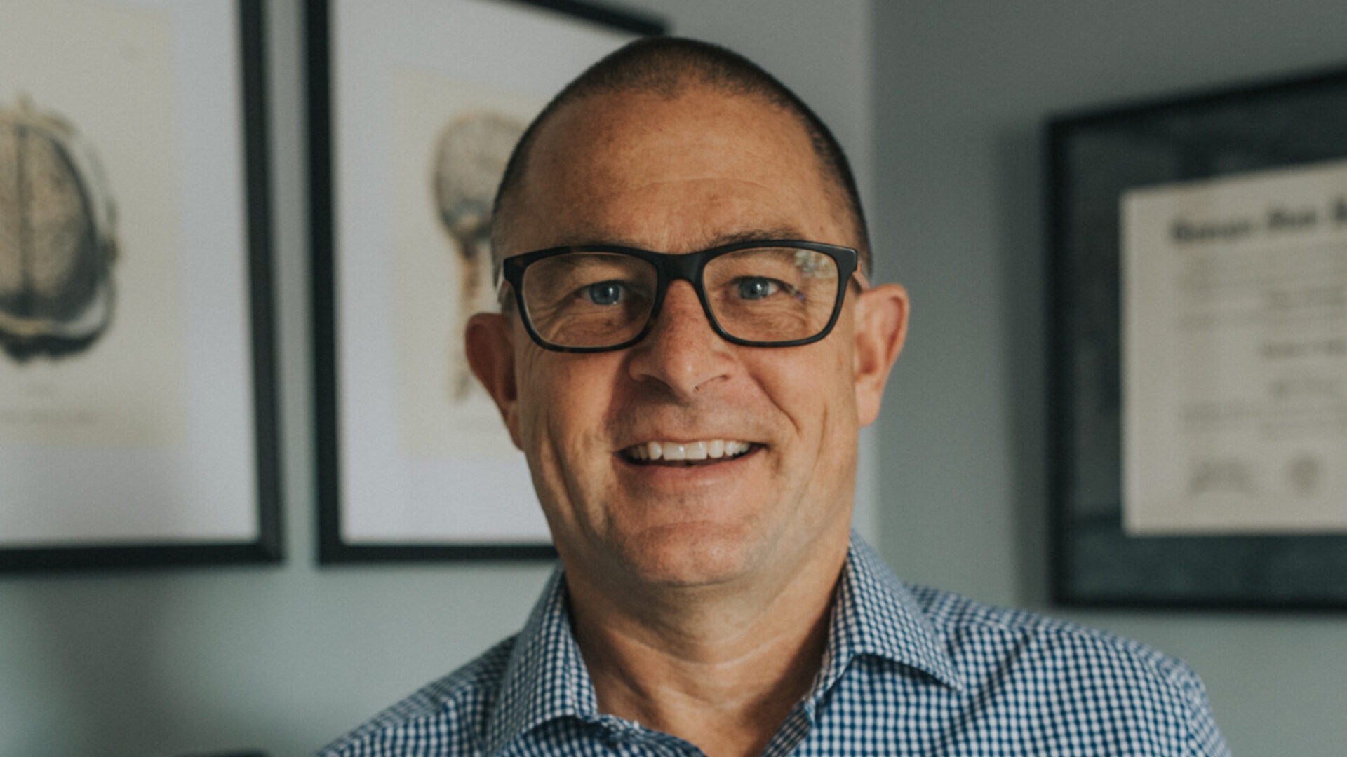 Man in checkered shirt holding a brain model, standing in an office with framed certificates on the wall.