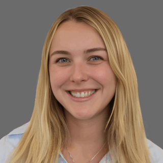 Smiling person with long blonde hair and a necklace, wearing a light blue collared shirt against a gray background.
