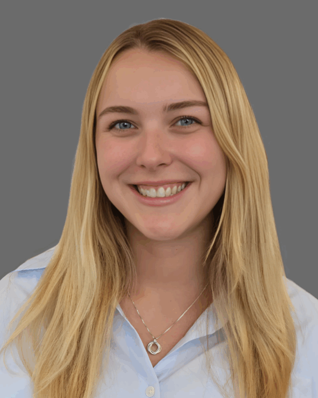 Smiling person with long blonde hair and a necklace, wearing a light blue collared shirt against a gray background.