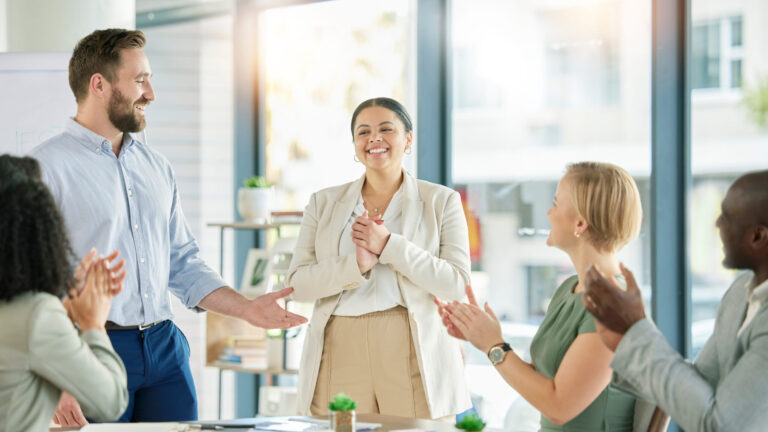 People clapping in an office meeting, smiling and congratulating a standing colleague