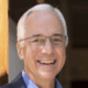 Smiling man with glasses and white hair in a blue shirt and blazer, standing outdoors near a wooden beam.