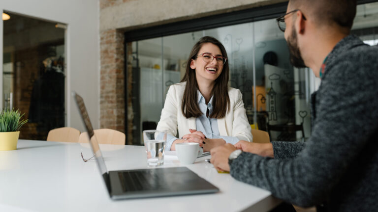 Two colleagues smiling and talking during a meeting in a modern office with a laptop and coffee on the table.
