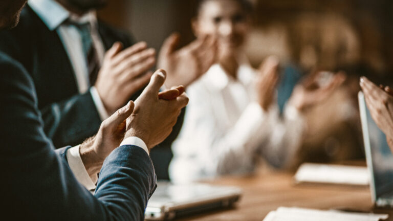 Group of people in business attire clapping during a meeting at a wooden table with papers and a smartphone.