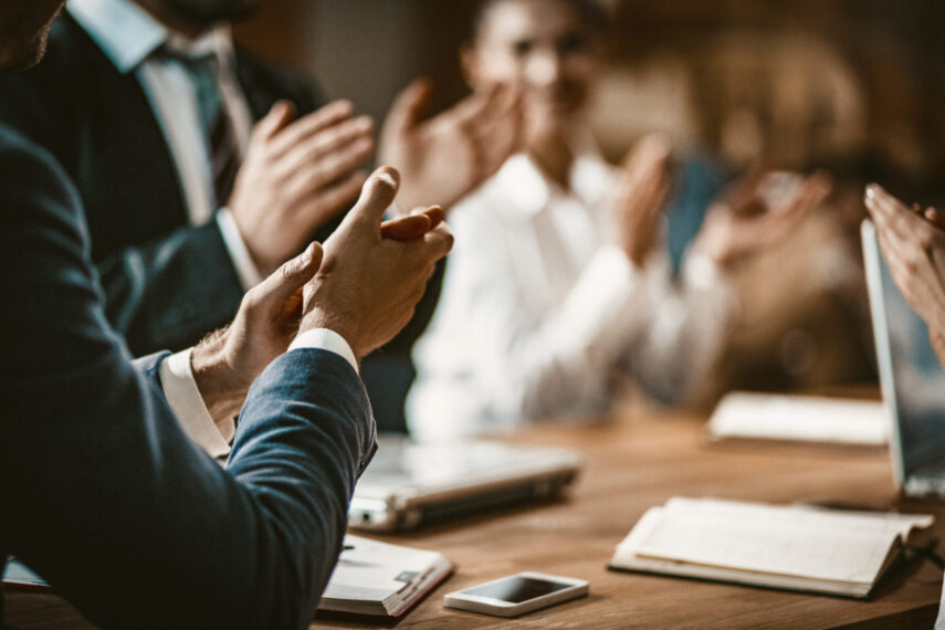 Group of people in business attire clapping during a meeting at a wooden table with papers and a smartphone.
