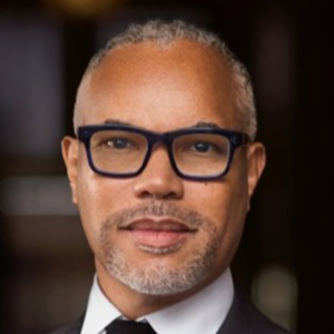 Man wearing glasses and a suit with a slight smile, set against a dark background.