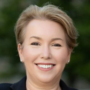 Smiling person with short hair, wearing a dark outfit, outdoors with greenery blurred in the background.