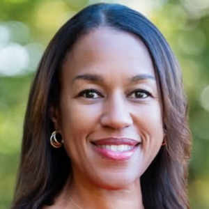 Smiling person with long hair and hoop earrings, standing outdoors with a blurred green background.