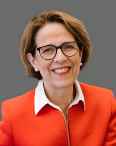 A smiling person in glasses and a red jacket with a white collar against a gray background.