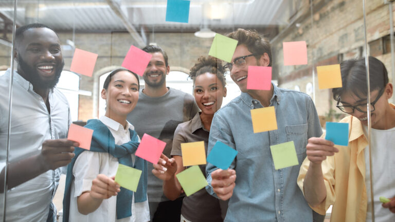 Diverse group of people smiling and placing colorful sticky notes on a glass wall in a bright office space.