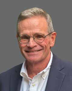 Smiling person with glasses wearing a white shirt and blazer against a gray background.