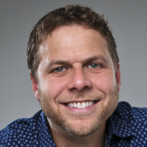 Smiling person wearing a patterned blue shirt against a gray background.