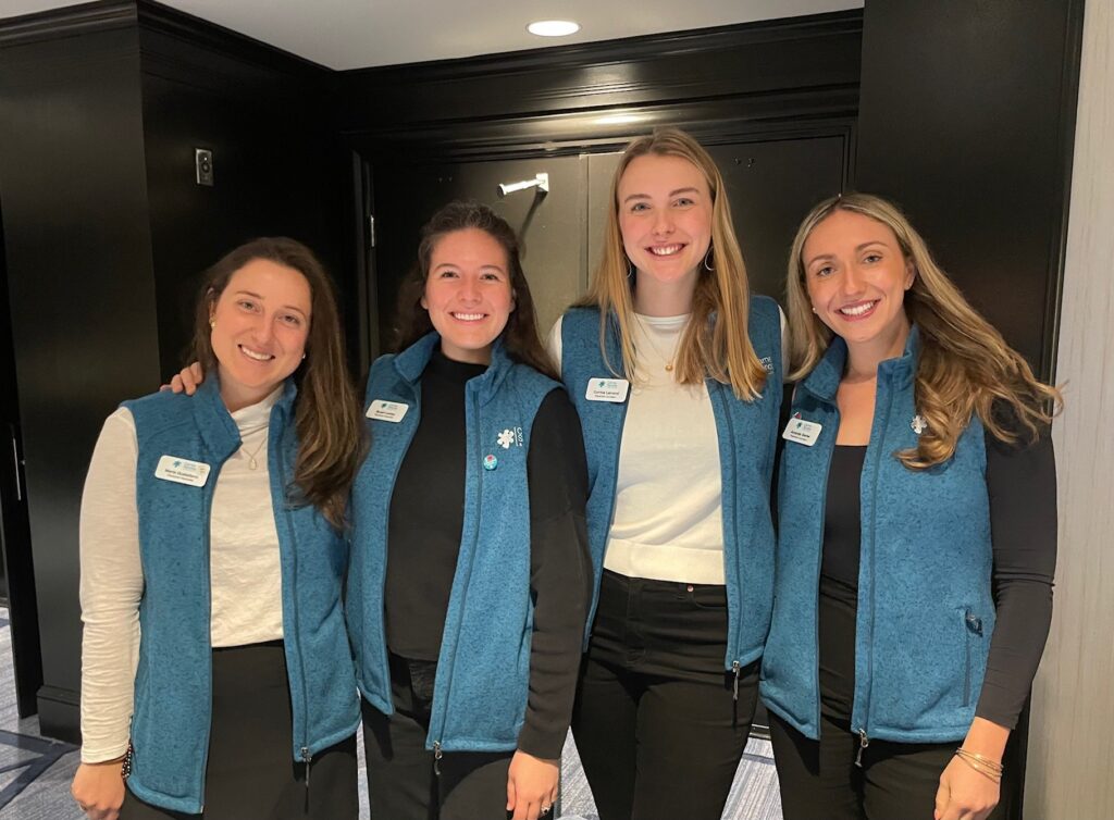 Four smiling people wearing blue vests standing together indoors, in front of a dark wooden wall.