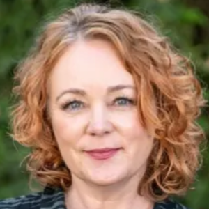 Person with curly hair smiling, posing outdoors with greenery in the background.