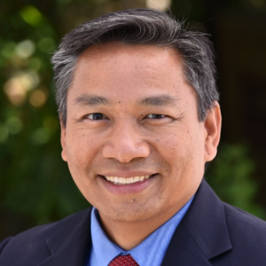 Smiling person in a suit and red tie standing outdoors with a blurred green background.