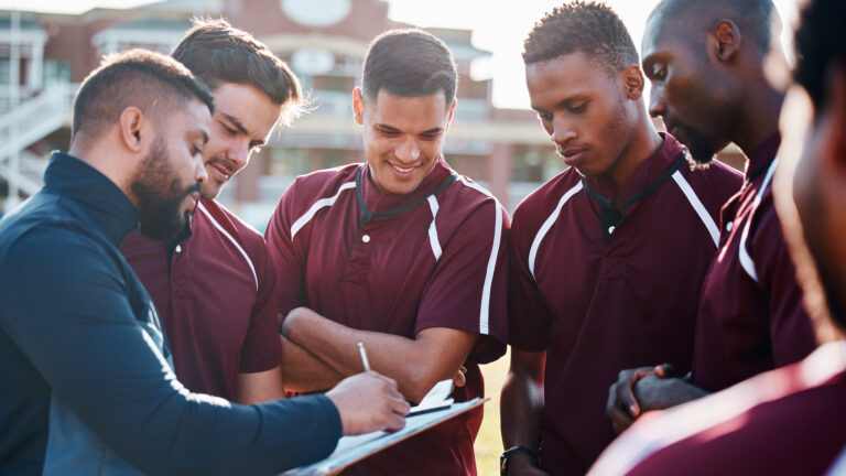 Coach discusses strategy with rugby team wearing maroon jerseys at the field.
