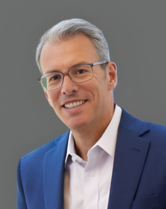 Smiling man in glasses wearing a blue suit jacket and white shirt against a neutral background.