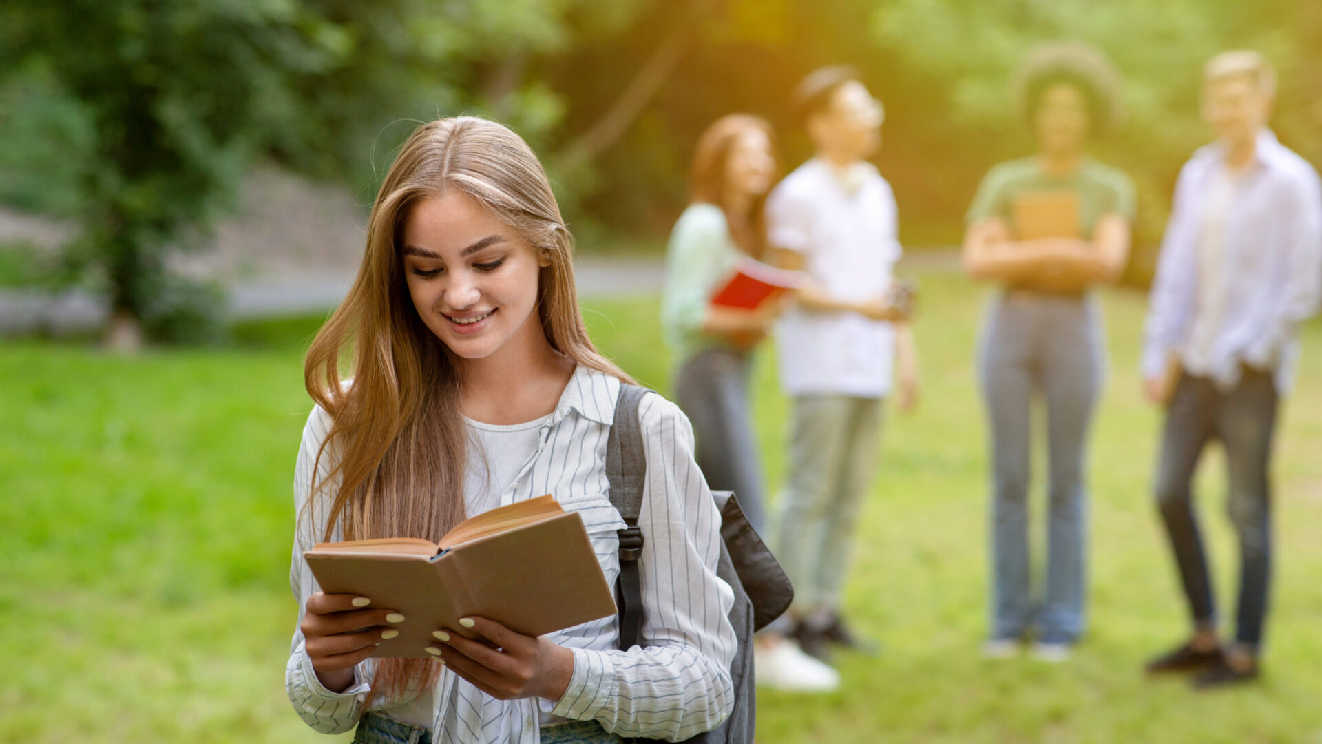 Young woman reading a book outdoors with a group of friends in the background on a sunny day.