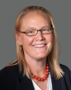 Person with glasses, smiling, wearing a black blazer and coral necklace against a gray background.