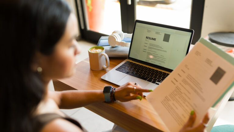 Woman reviewing a resume on paper and laptop at a wooden table in a cafe.