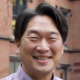 Smiling person standing outdoors in front of brick building.