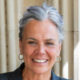 Smiling person with gray hair and wearing hoop earrings in front of a stone column background.