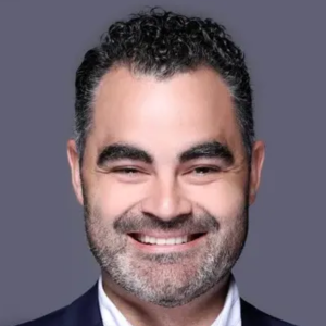 Smiling man with short curly hair and beard, wearing a dark suit jacket against a gray background.