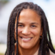 Smiling person with long braided hair, wearing a brown top, with a blurred background.