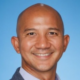 Smiling bald man with blue background wearing a suit jacket.