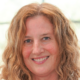 Woman with curly hair smiling warmly at the camera, with a blurred indoor background.